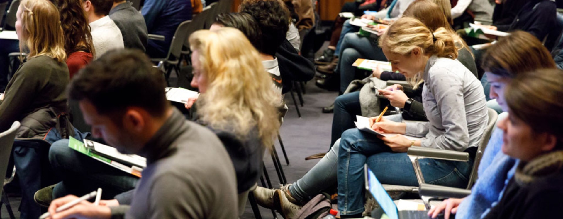 Viele sitzende Menschen in einem Konferenzsaal, die dem Vortrag zuhören oder an digitalen Endgeräten arbeiten.