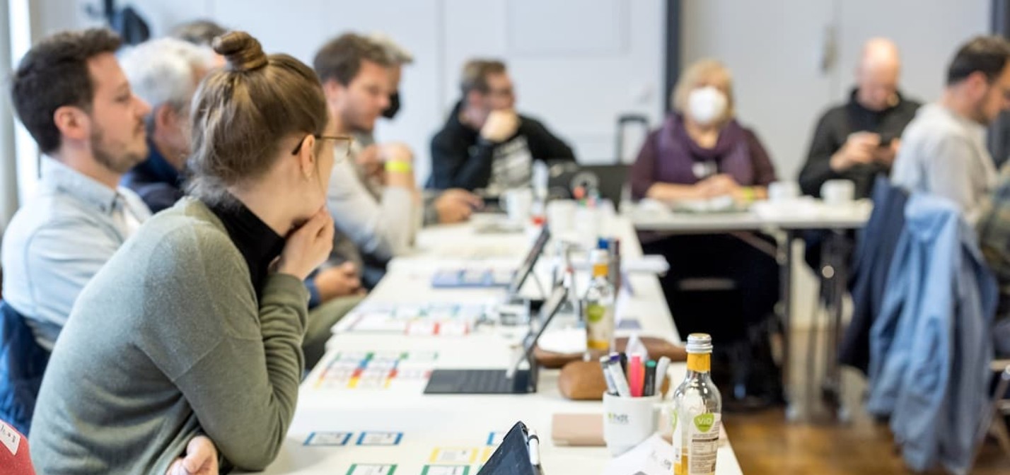 Menschen, die an Tischen sitzen und zuhören. Auf den Tischen liegen Tablet und farbige Karten.